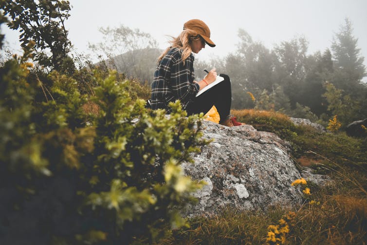 A woman  sitting outdoors writing in a journal.